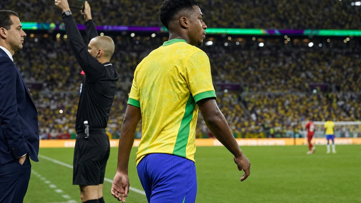 Jogador jovem de futebol entrando em campo sob luzes de estádio durante partida internacional.