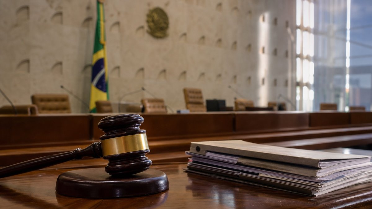 Plenário judicial vazio com documentos, martelo e bandeira do Brasil ao fundo.