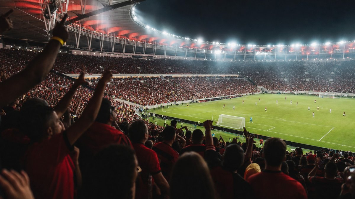 Torcedores em arquibancada de estádio brasileiro acompanhando uma partida de futebol à noite.