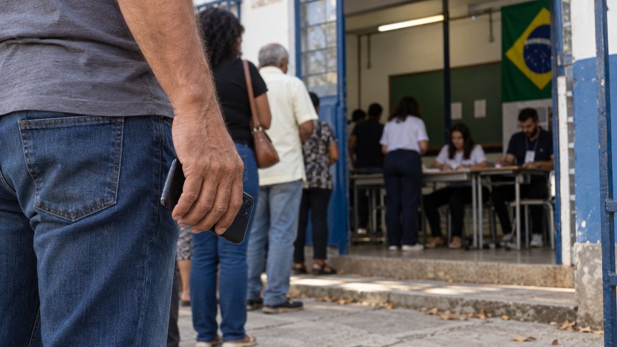 Entrada de local de votação em Minas Gerais com eleitor segurando celular fora da cabine.